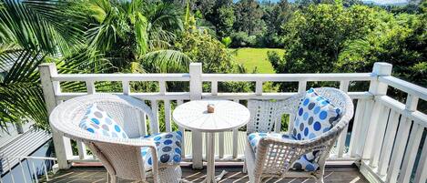 Une terrasse en bois avec deux fauteuils en osier et une petite table ronde, offrant une vue sur un paysage verdoyant et luxuriant, avec des arbres et une chaîne de montagnes au loin.