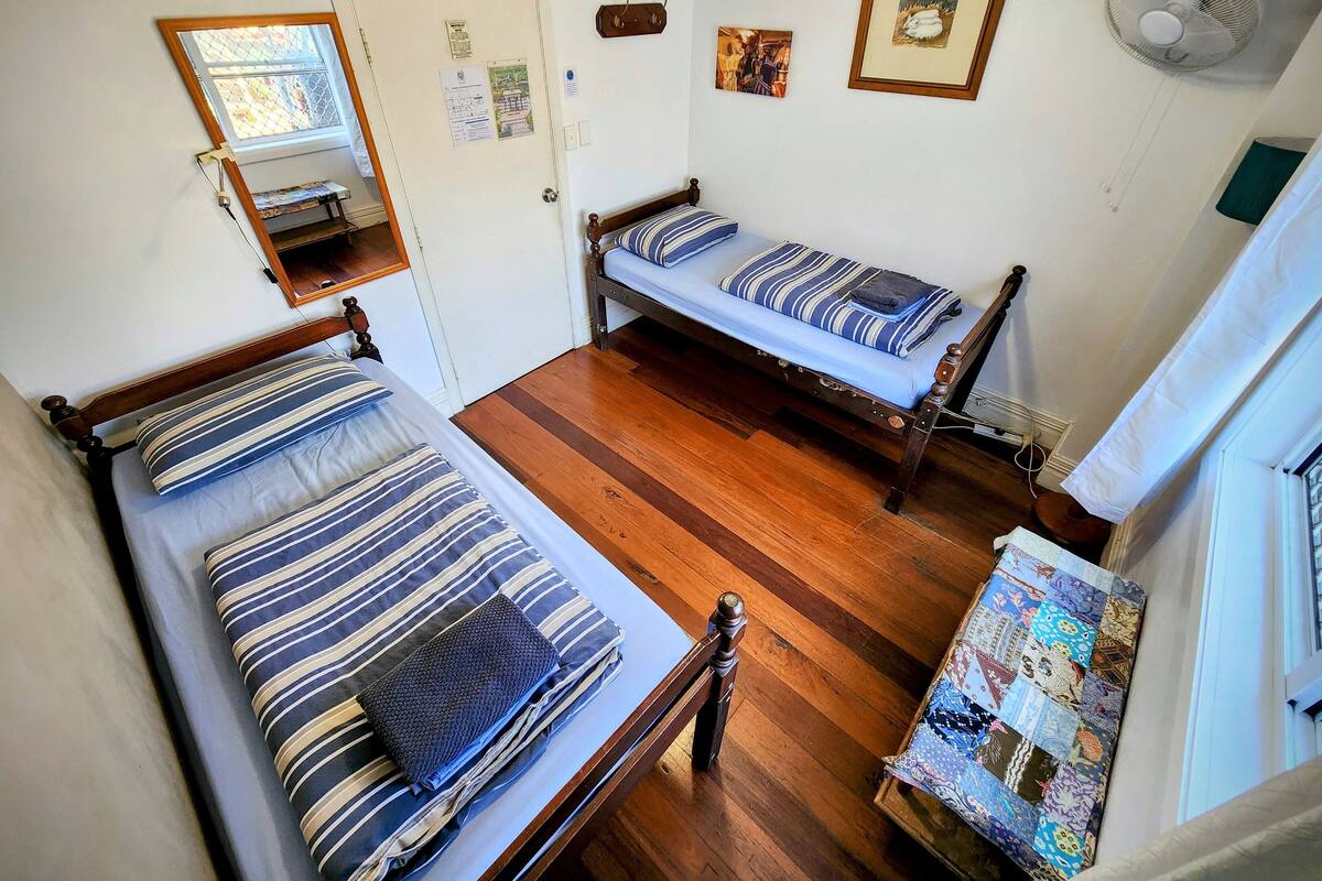 Ein Zimmer mit zwei hölzernen Betten, einem kleinen Fenster, einer Tür und einem Wandventilator.