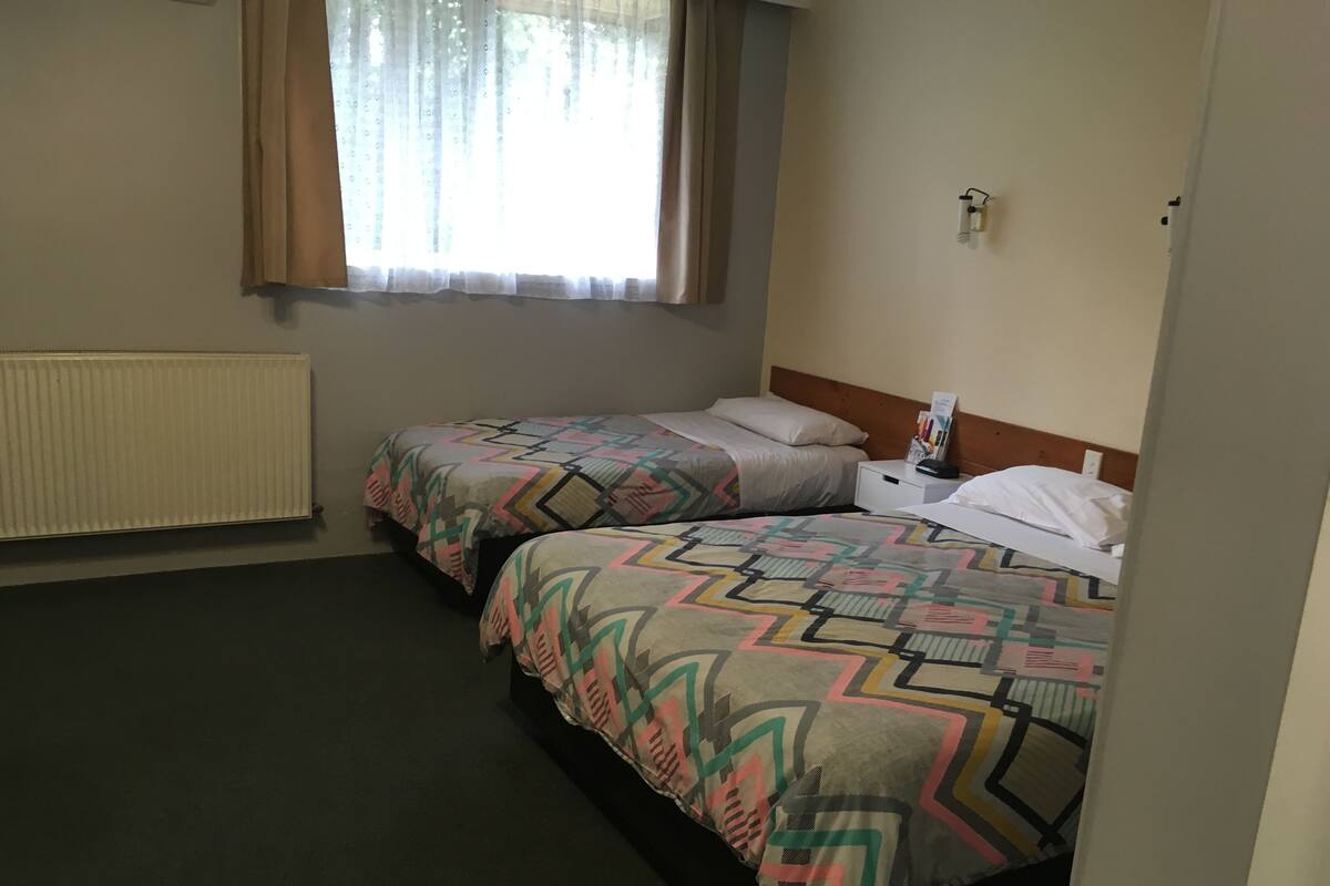 A hotel room with two beds, a window with curtains, a radiator, and a nightstand with a lamp.