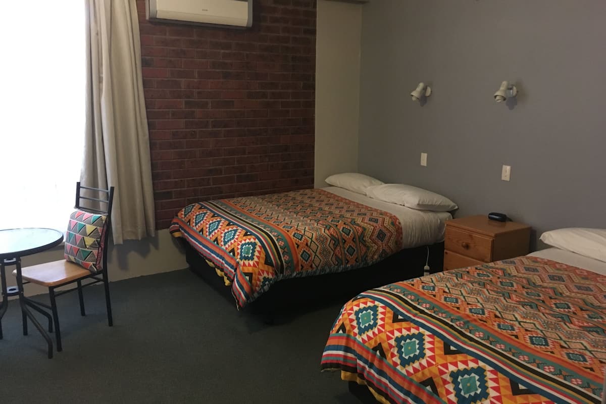 A hotel room with two beds, a small table, a chair, and a brick wall.