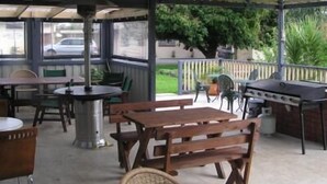 Eine Terrassengruppe im Freien mit Holztischen und -stühlen, einem Grill und einem überdachten Bereich mit Blick auf die Straße und Grünflächen.