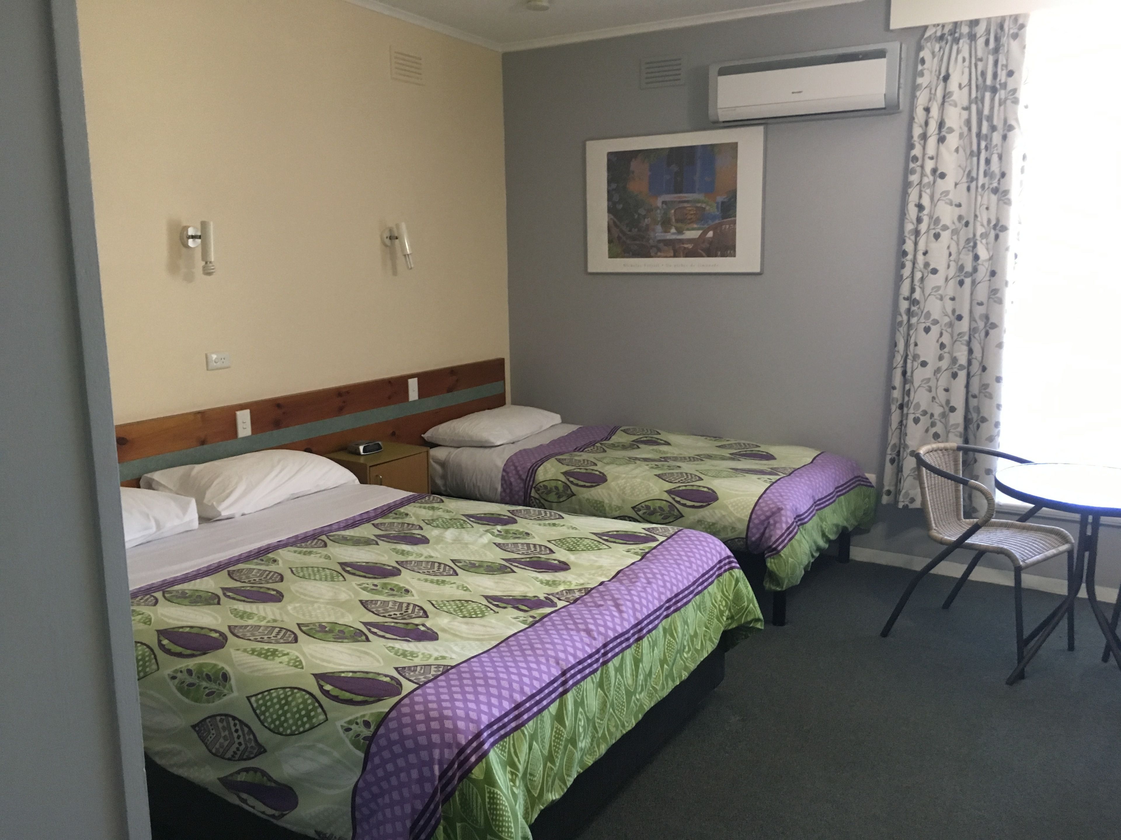 A hotel room with two beds, a small table, and a chair. There is a wall-mounted air conditioner and a framed picture above the bed.