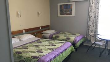 A hotel room with two beds, a small table, and a chair. There is a wall-mounted air conditioner and a framed picture above the bed.