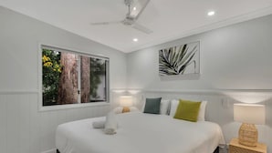 A bedroom with a large bed, a ceiling fan, a window with a view of trees, and a wall-mounted plant.
