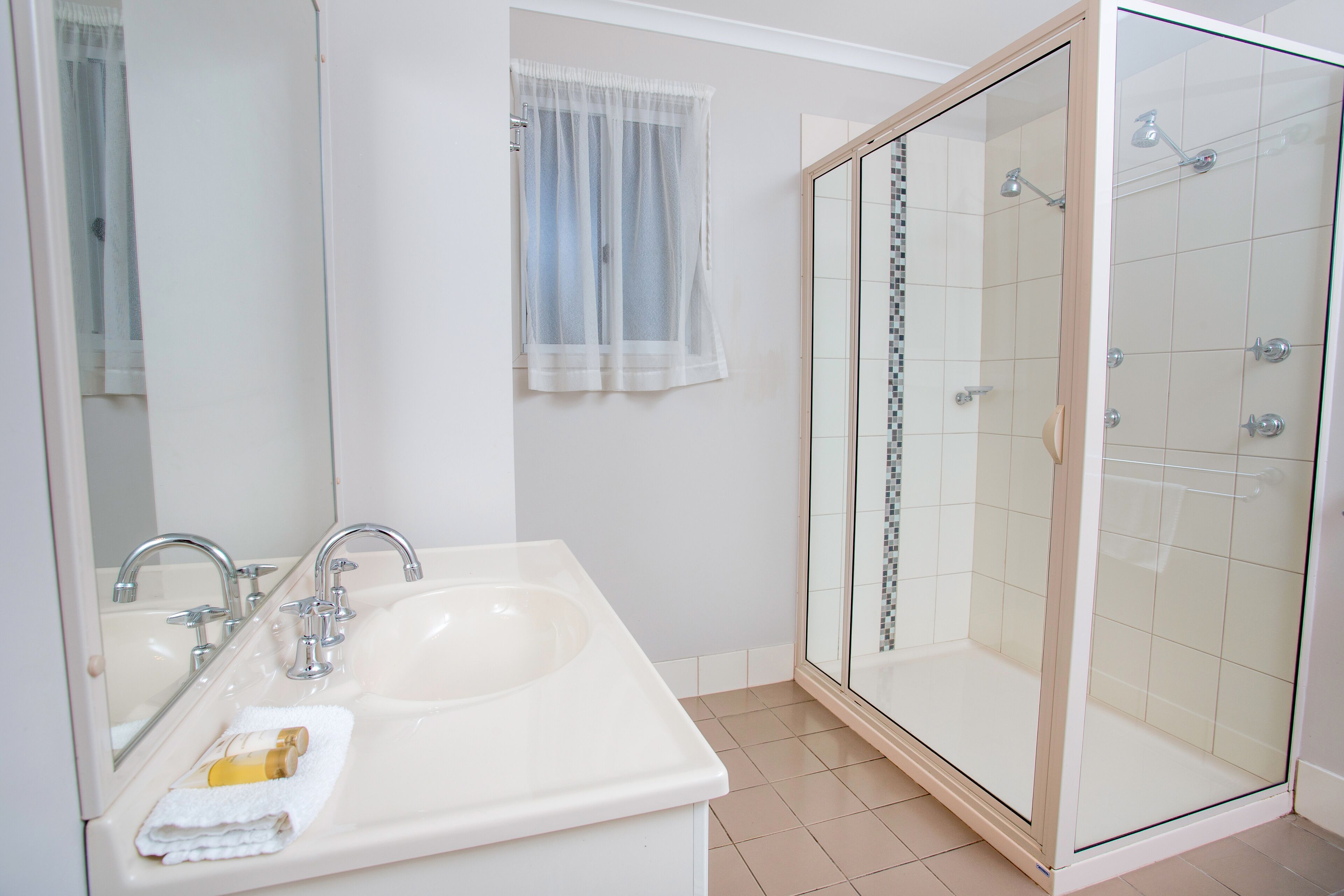 A bathroom with a double sink, a glass shower enclosure, and a window with sheer curtains.