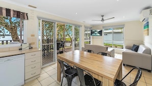 A modern kitchen with a dishwasher, a dining area with a wooden table and chairs, a sofa, and a sliding glass door leading to a balcony with a view of trees and a body of water.