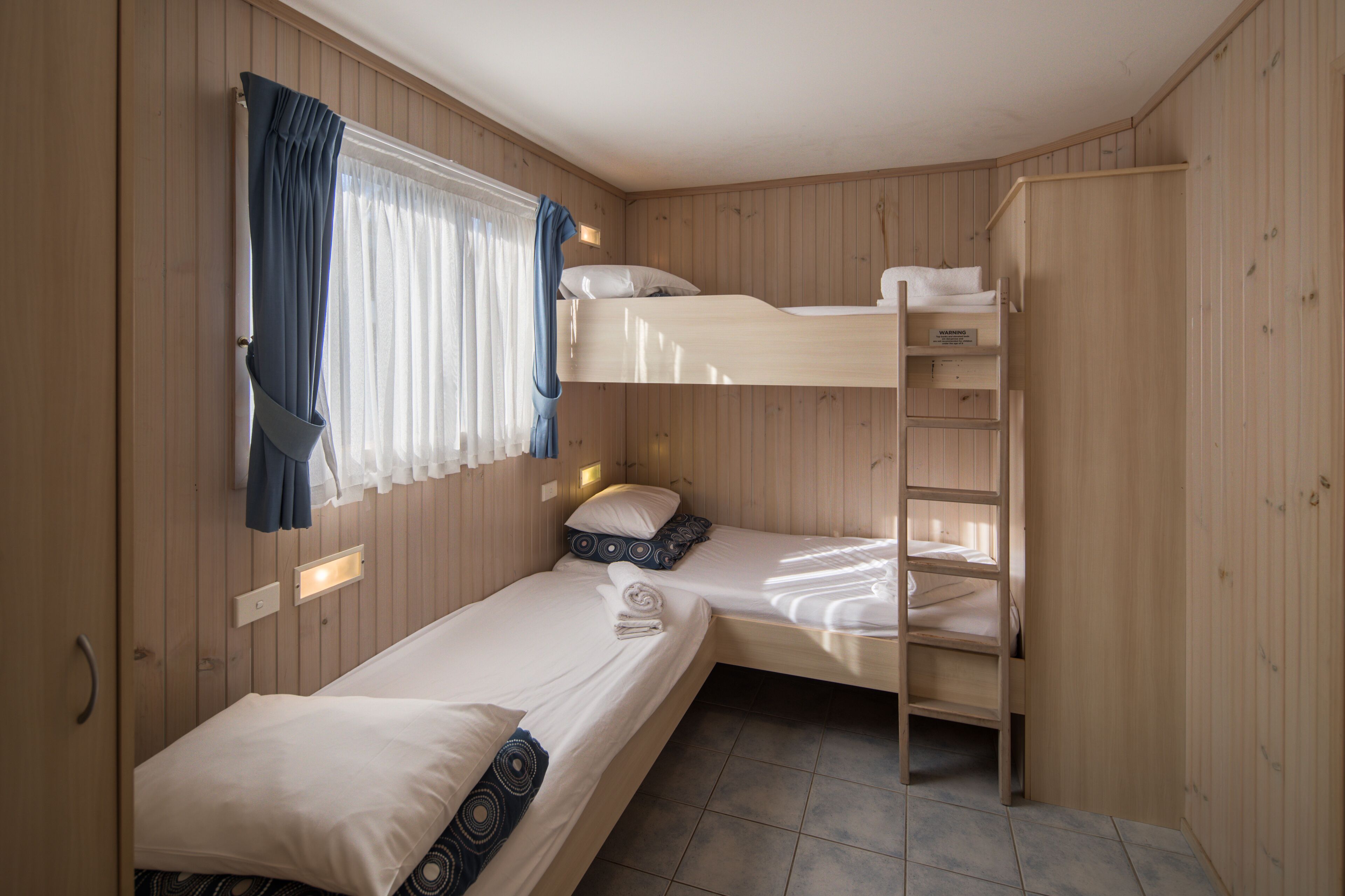 Ein Schlafraum mit Etagenbett, Holzwänden, einem Fenster mit Vorhängen und einem fest installierten Regal.