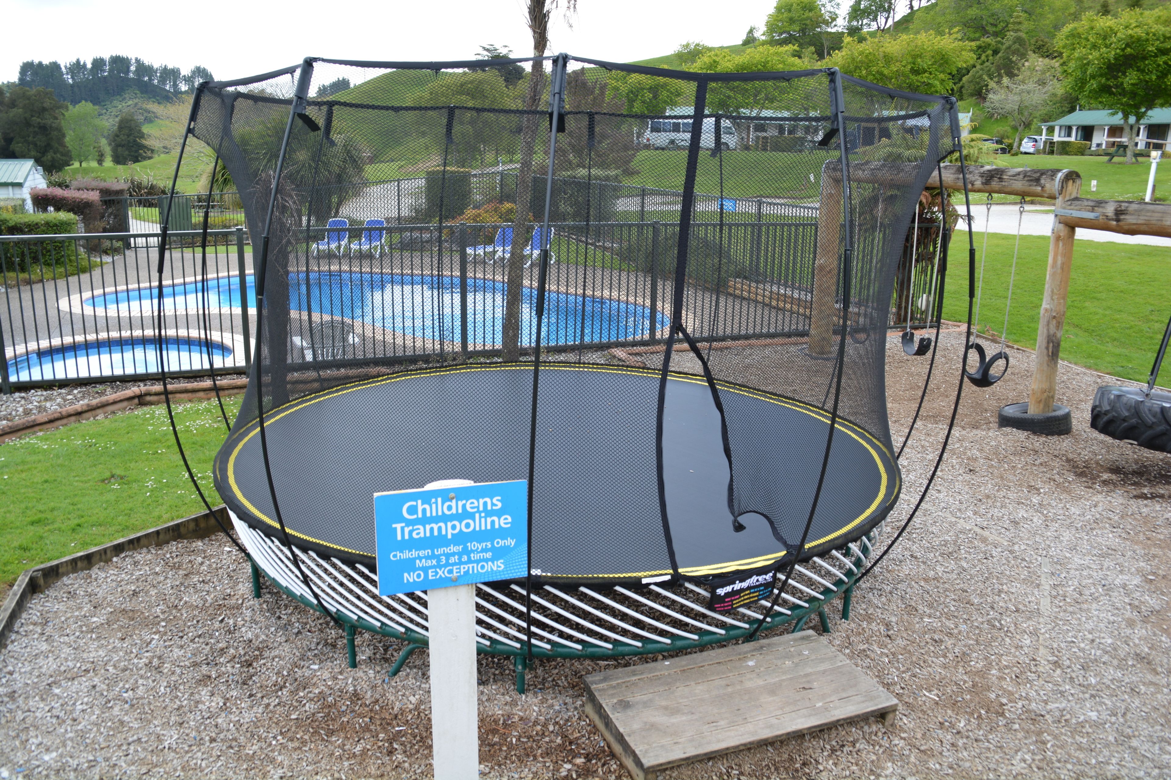 A children's trampoline with a safety net, surrounded by a grassy area and a pool.