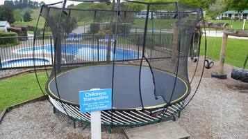 A children's trampoline with a safety net, surrounded by a grassy area and a pool.