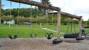Un ensemble de balançoires en bois, avec des sièges en forme de pneu, installé dans un espace aménagé en parc, avec un sol en gravier, entouré de verdure et bordé d’un sentier.