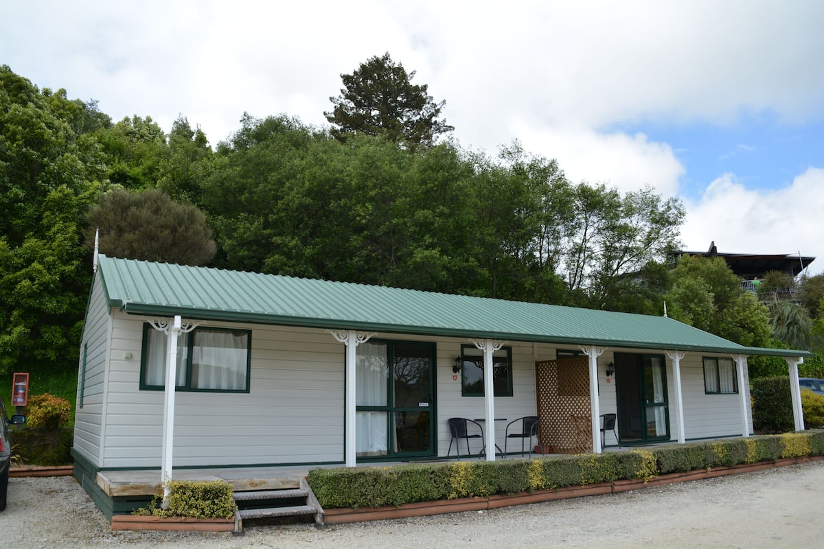 A single-story building with a green roof, a porch with two chairs, and a small front garden.