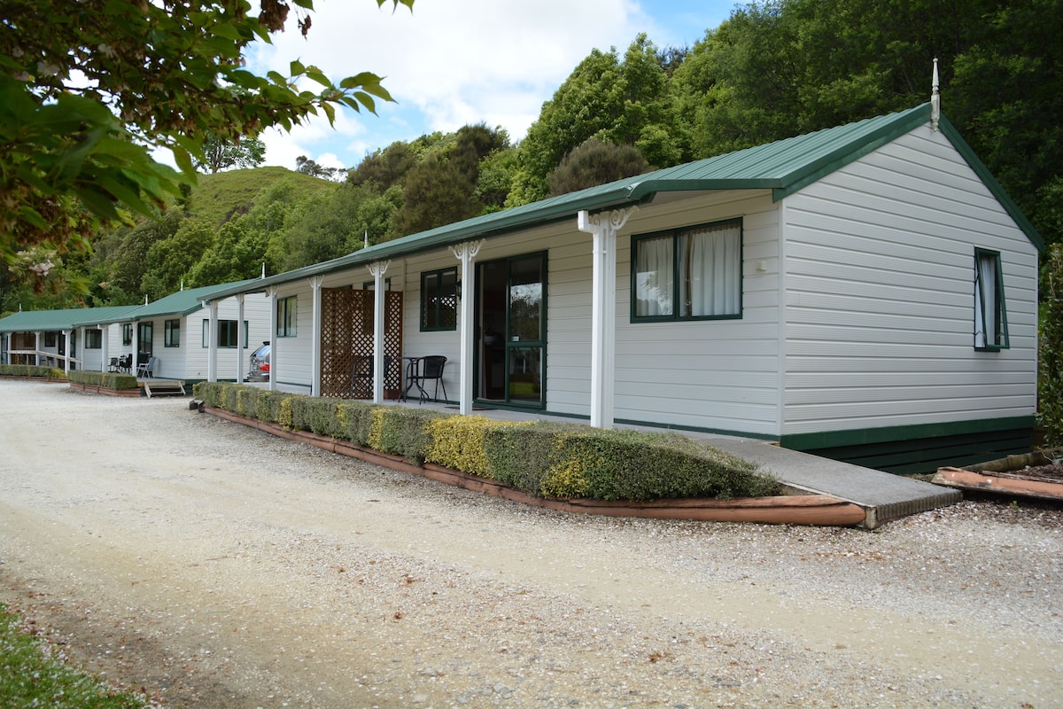A row of white mobile homes with green roofs, each with a porch and a small table.