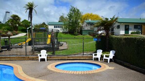 Une piscine bordée de briques, entourée de chaises blanches et d’une aire de jeux clôturée.