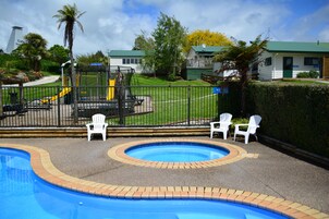 Piscina com borda de tijolos, rodeada por cadeiras brancas e uma área de recreação cercada.