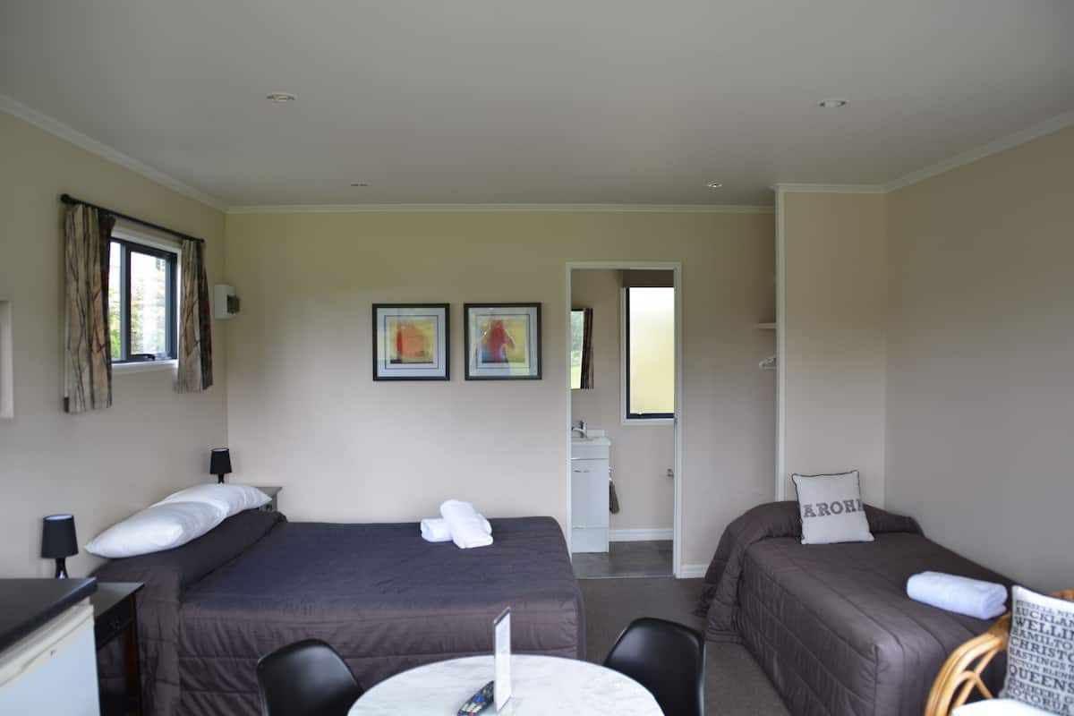 A hotel room with two beds, a desk, a chair, a small table, and a bathroom visible through an open door.
