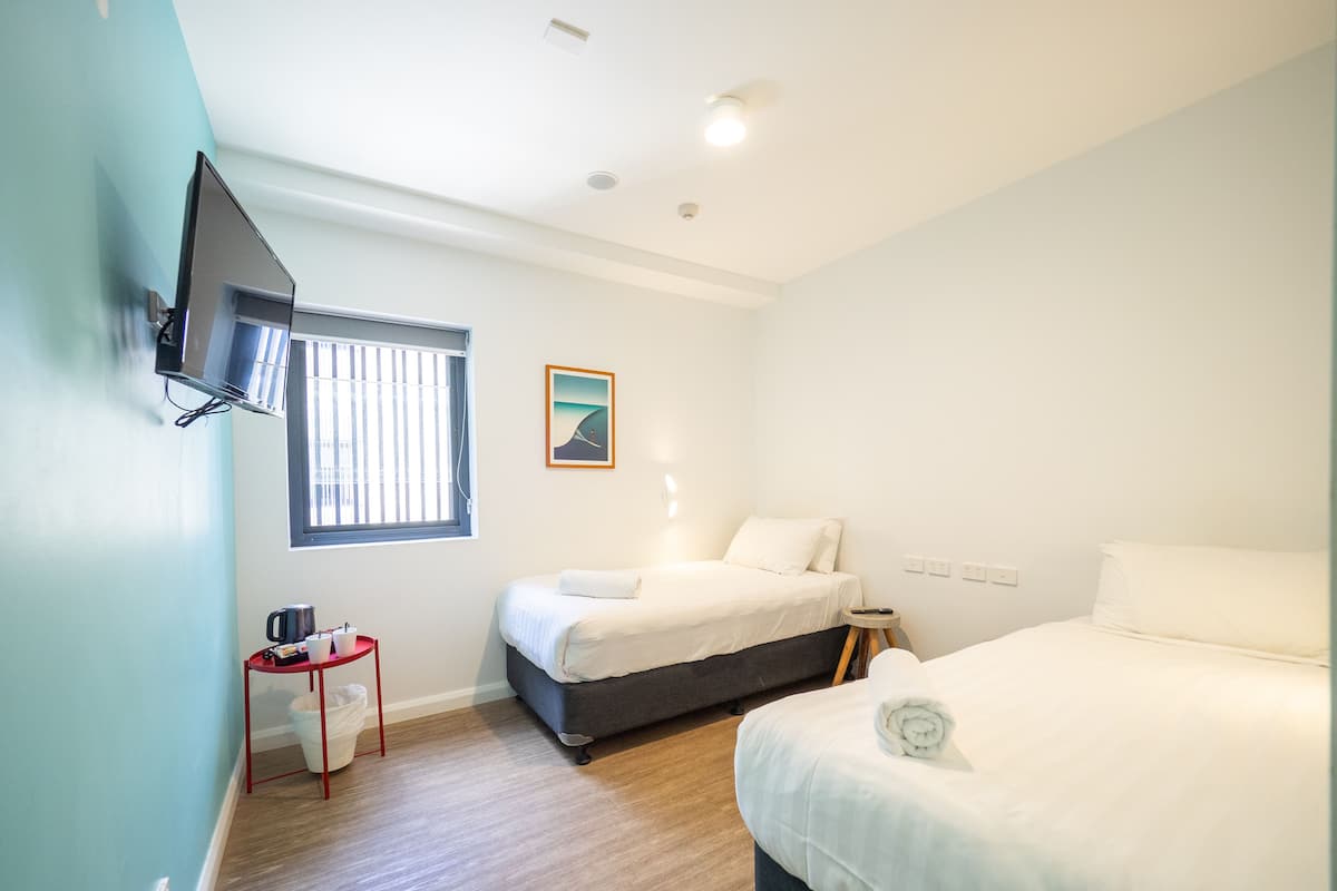 A hotel room with two beds, a TV mounted on the wall, a small table with a kettle, and a window with blinds.