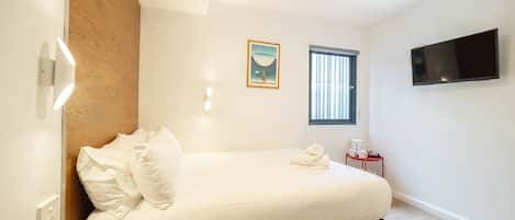 A bedroom with a bed, a bedside table with a cup and a book, a window with blinds, a painting on the wall, and a TV mounted on the wall.
