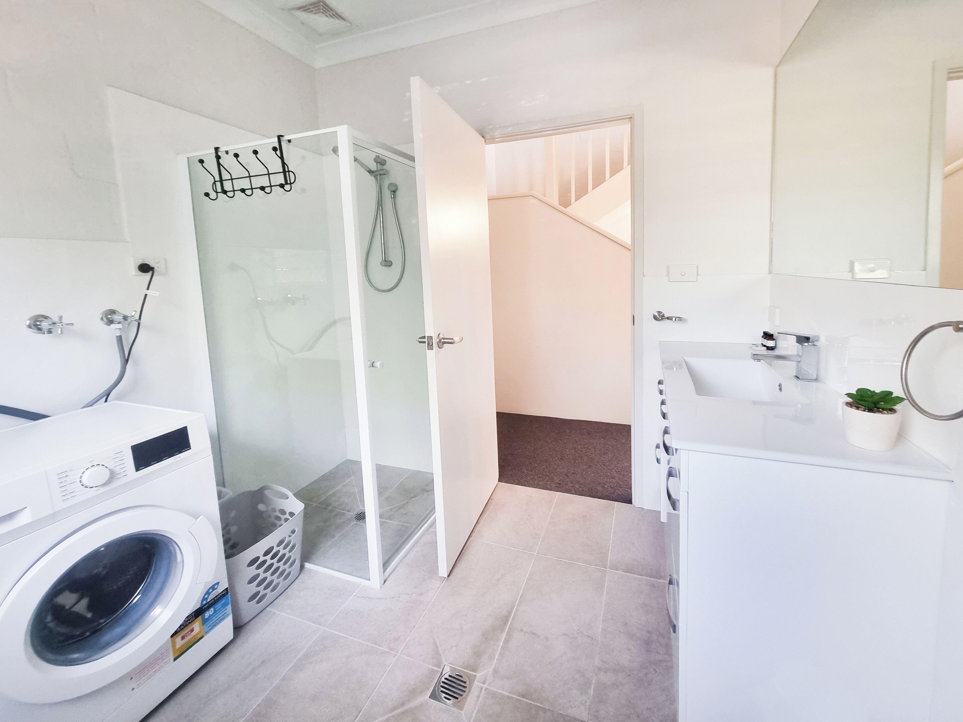 A modern bathroom with a glass shower enclosure, a washing machine, a sink, and a small potted plant.