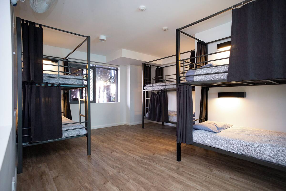 A room with bunk beds, wooden flooring, and a window with curtains.