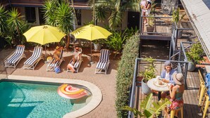 Zona de piscina con sillas de descanso y sombrillas, rodeada de palmeras y plantas en macetas.