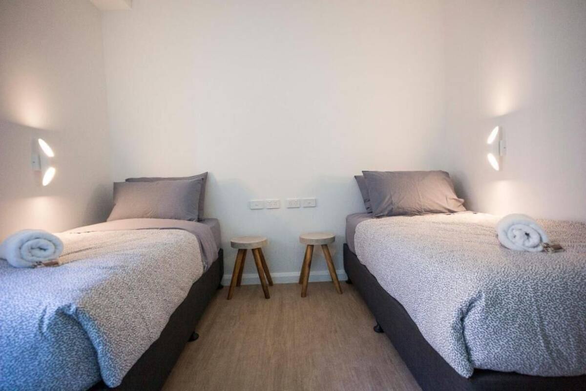A room with two beds, each with a pillow and a rolled towel, a small stool between them, and a light switch on the wall.