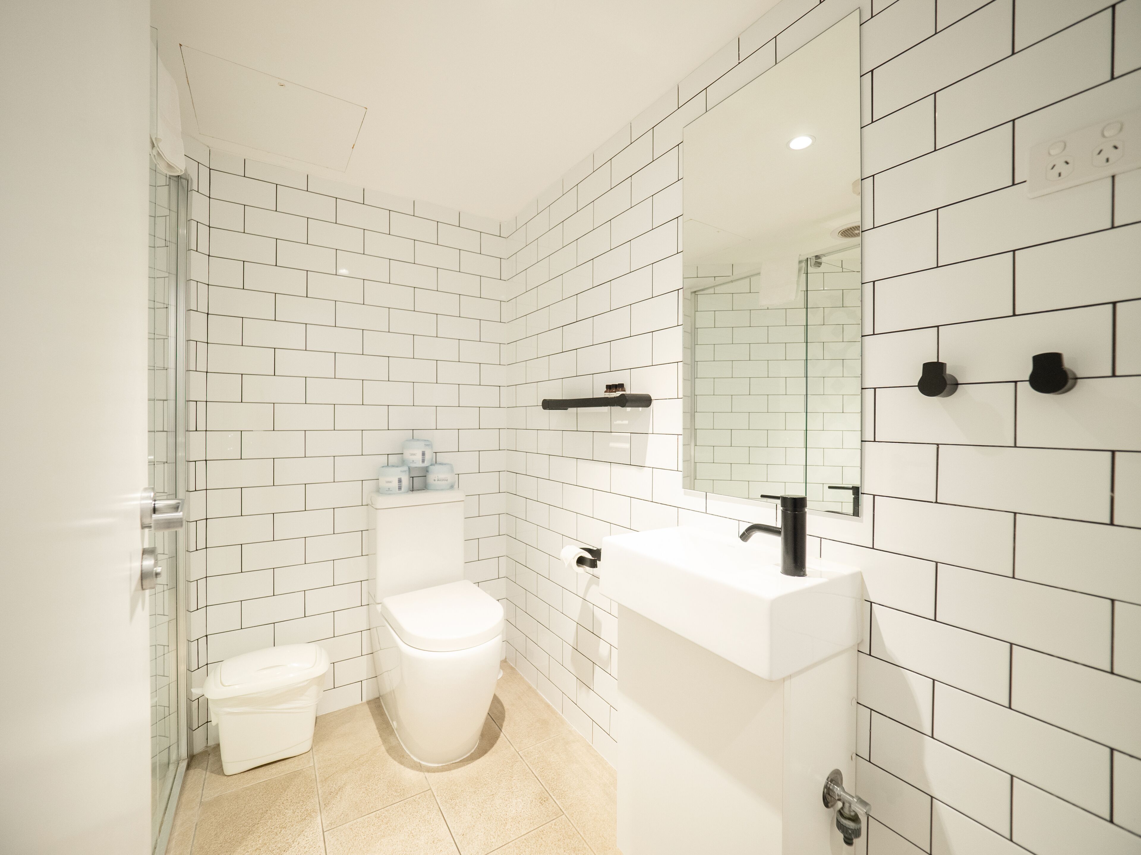 A modern bathroom with white subway tiles, a white toilet, a white sink with a black faucet, and a mirror above the sink.
