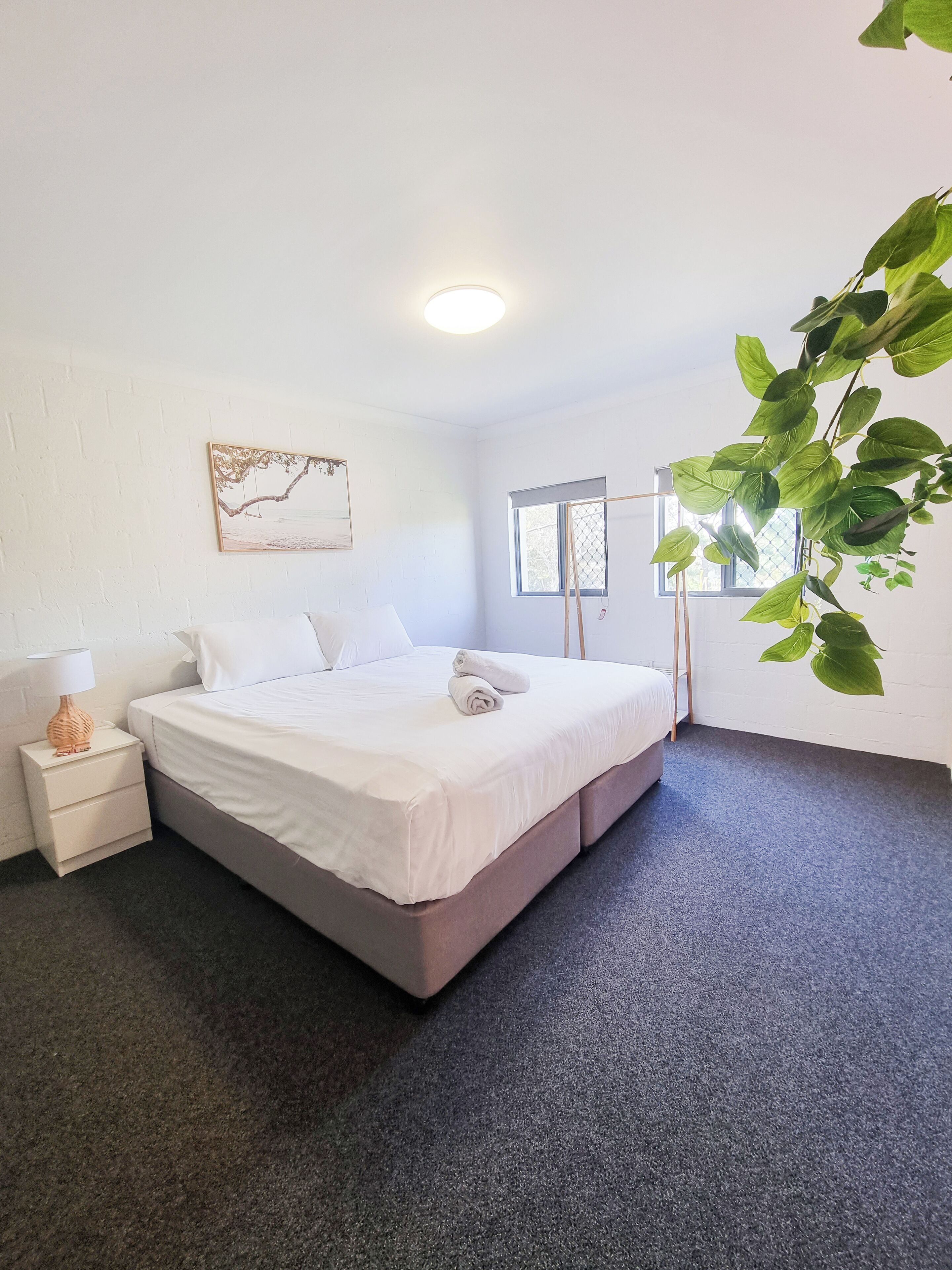 A neatly made bed with a white comforter and pillows, a bedside table with a lamp, a framed picture on the wall, and a window with a view of trees.