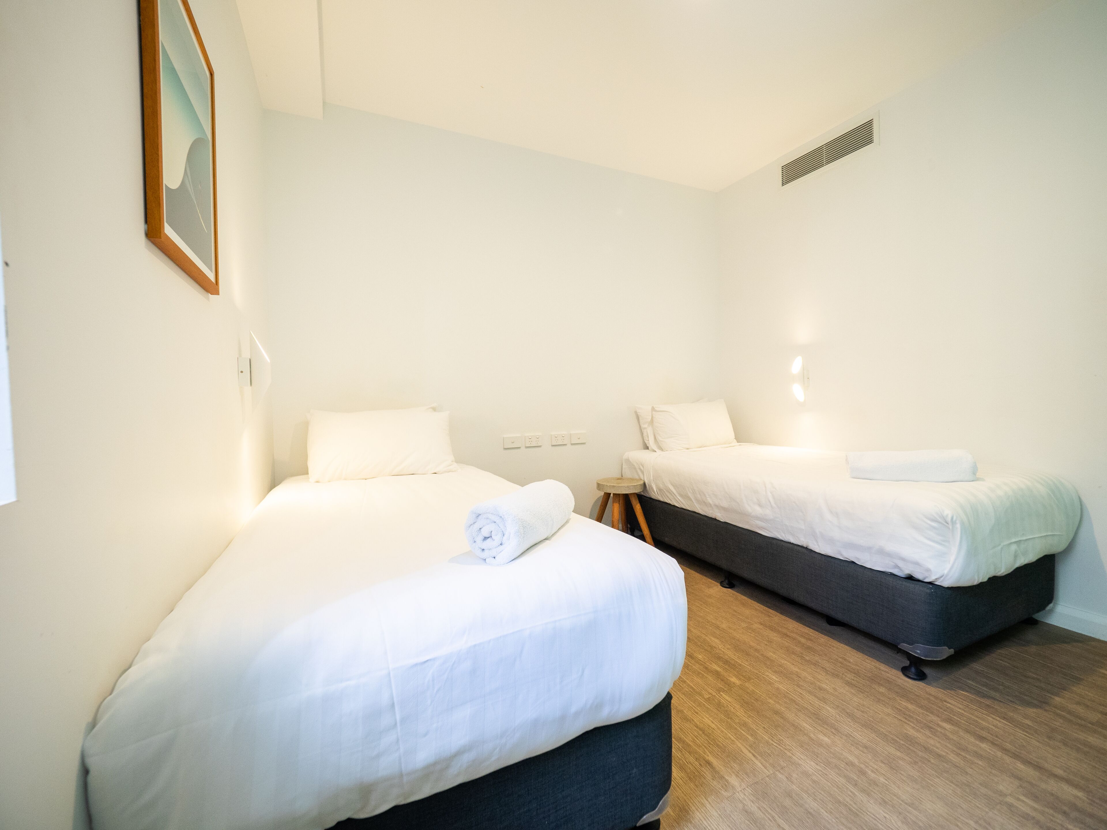 A hotel room with two beds, a small table, and a painting on the wall.
