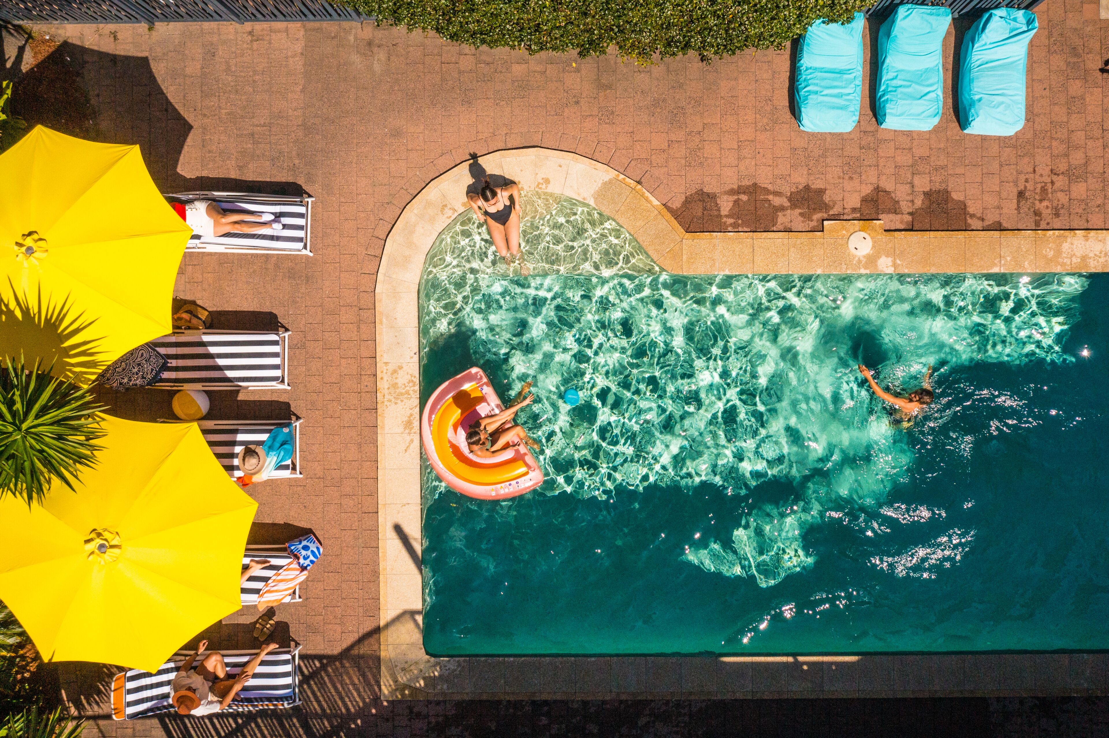 An aerial view of a swimming pool with lounge chairs and umbrellas, surrounded by a brick patio.