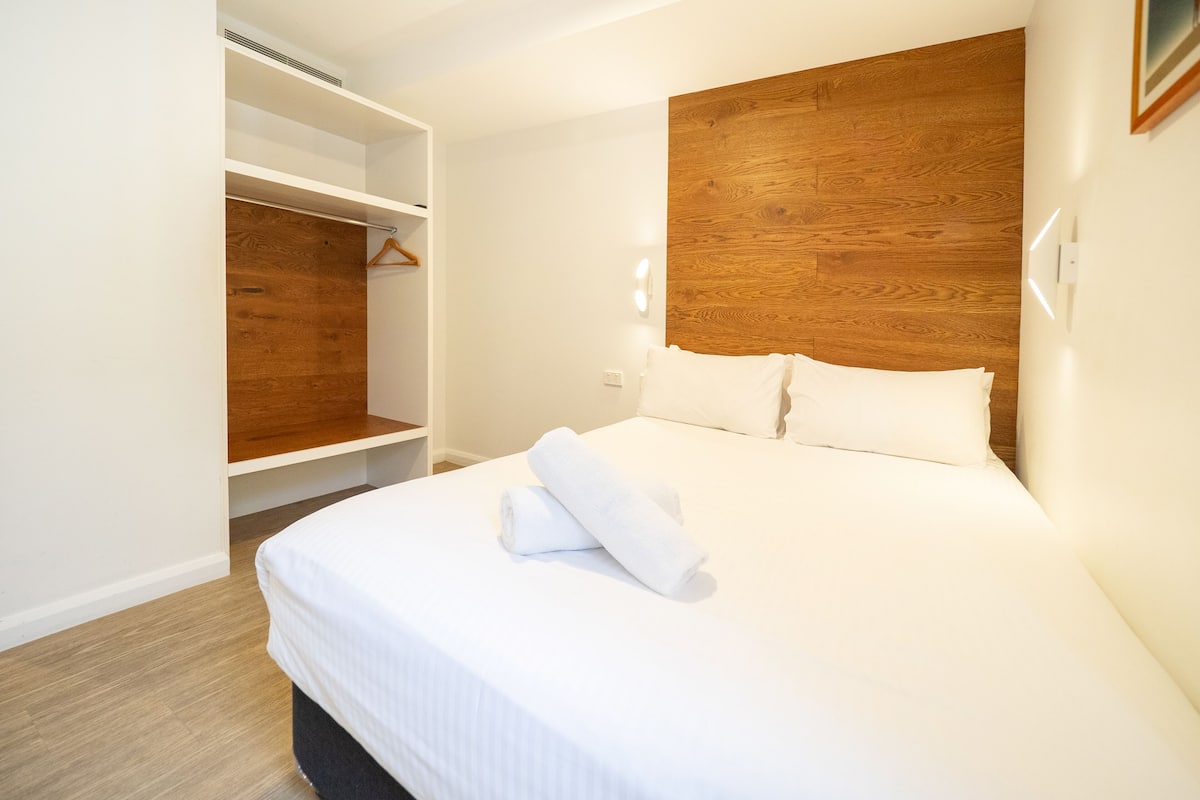 A hotel room with a bed, a wooden headboard, a wardrobe with a hanging rack, and a built-in heater.