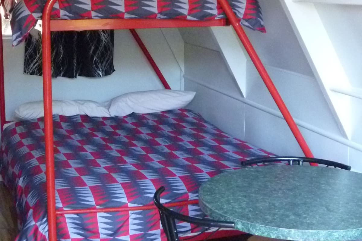 A bunk bed with a patterned bedspread, a small table with chairs, and a window with curtains.