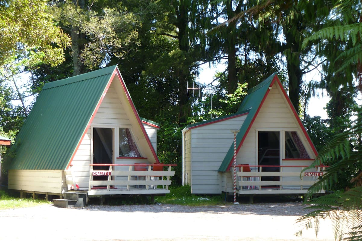Two small, triangular-roofed cabins with a porch, labeled "CHALET 1" and "CHALET 2", surrounded by dense forest.