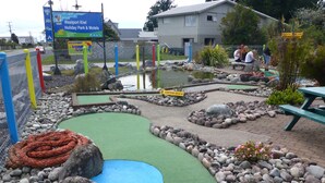 Un parcours de minigolf avec un tapis vert pour les putts, entouré de rochers et de galets, et une enseigne indiquant le « Westport Kiwi Holiday Park & Motels » en arrière-plan.