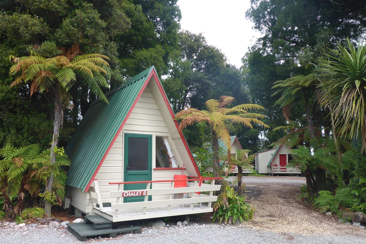 A small, single-story chalet with a green roof and white walls, surrounded by lush greenery.