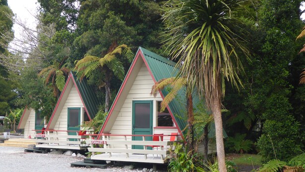 Deux cabanes de style chalet, avec des boiseries vertes et des touches de rouge, entourées d’une végétation luxuriante et posées sur un sol de gravier.