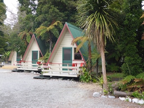 Dos cabañas de estilo chalet con detalles en verde y toques rojos, rodeadas de abundante vegetación y suelo de grava.