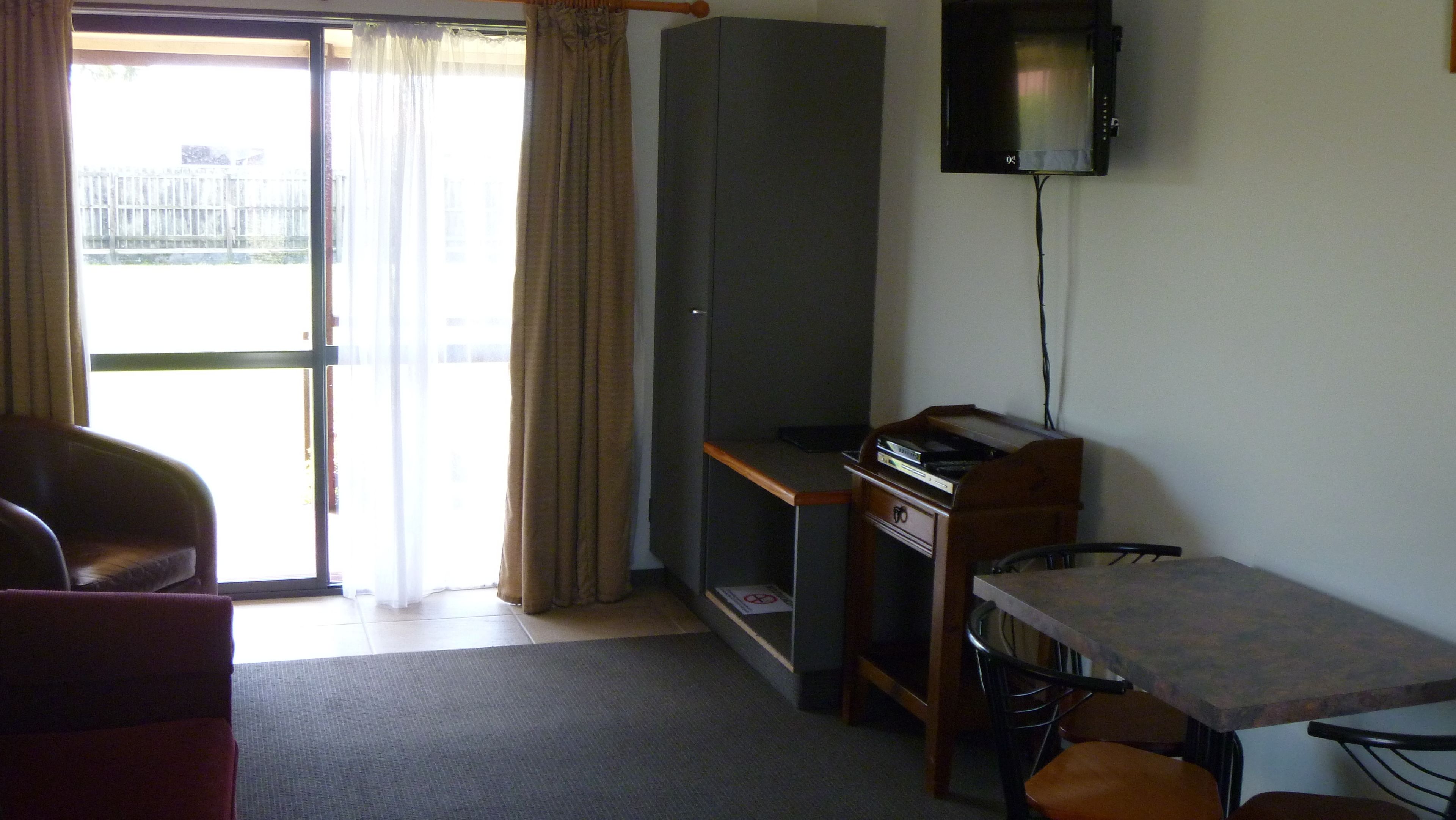 A room with a sliding glass door leading to a balcony, a small desk with a chair, a television mounted on the wall, and a sofa.