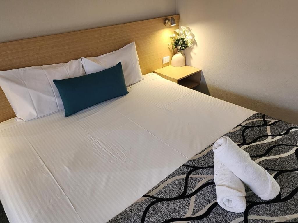 A neatly made bed with white linens, a teal pillow, and a towel on a patterned rug.