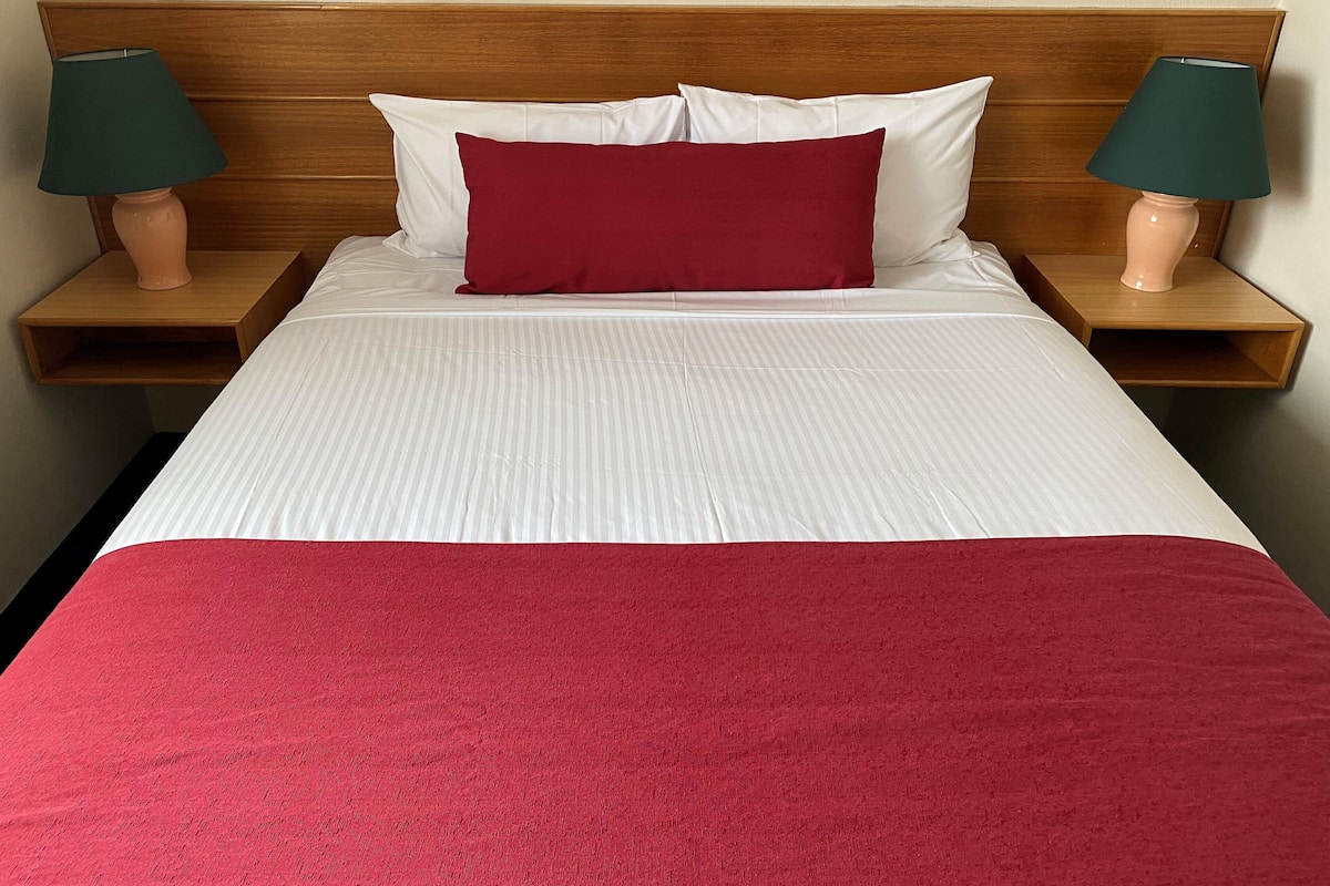 A neatly made bed with white bedding and a red throw blanket, flanked by two wooden nightstands with lamps.