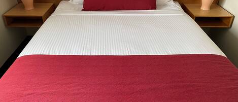 Una cama bien hecha con ropa blanca y una manta roja, flanqueada por dos mesitas de noche de madera con lámparas.
