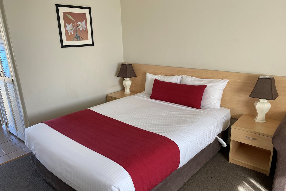 A hotel room with a bed, two bedside tables, a nightstand, a lamp, a framed picture on the wall, and a window with blinds.