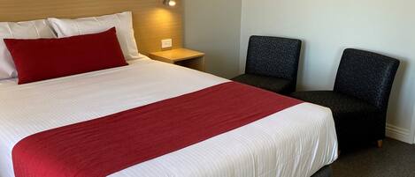 Habitación de hotel con una cama grande, una mesita de noche, dos sillas y una lámpara fijada a la pared.