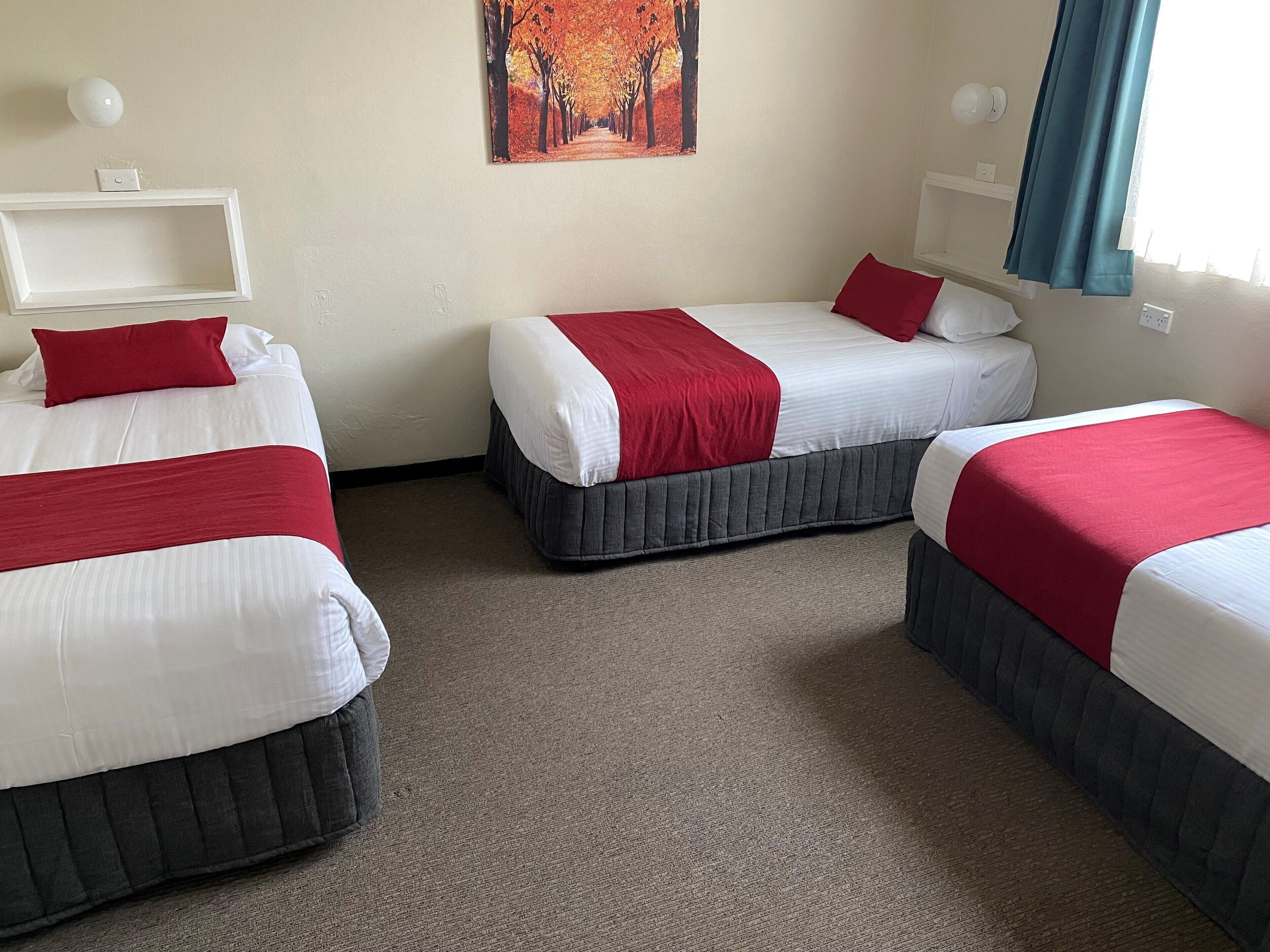Quarto de hotel com três camas, cada uma com roupa de cama branca e detalhes vermelhos. Há uma pintura de árvores na parede, uma estante branca e uma janela com cortinas.