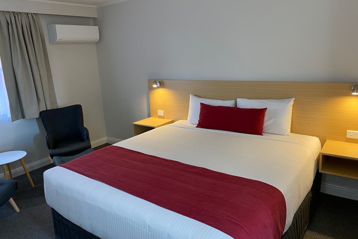 A hotel room with a large bed, a bedside table, a chair, a small table, and an air conditioning unit.