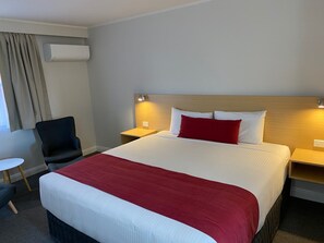 A hotel room with a large bed, a bedside table, a chair, a small table, and an air conditioning unit. - Country Gardens Motel (Coonabarabran)