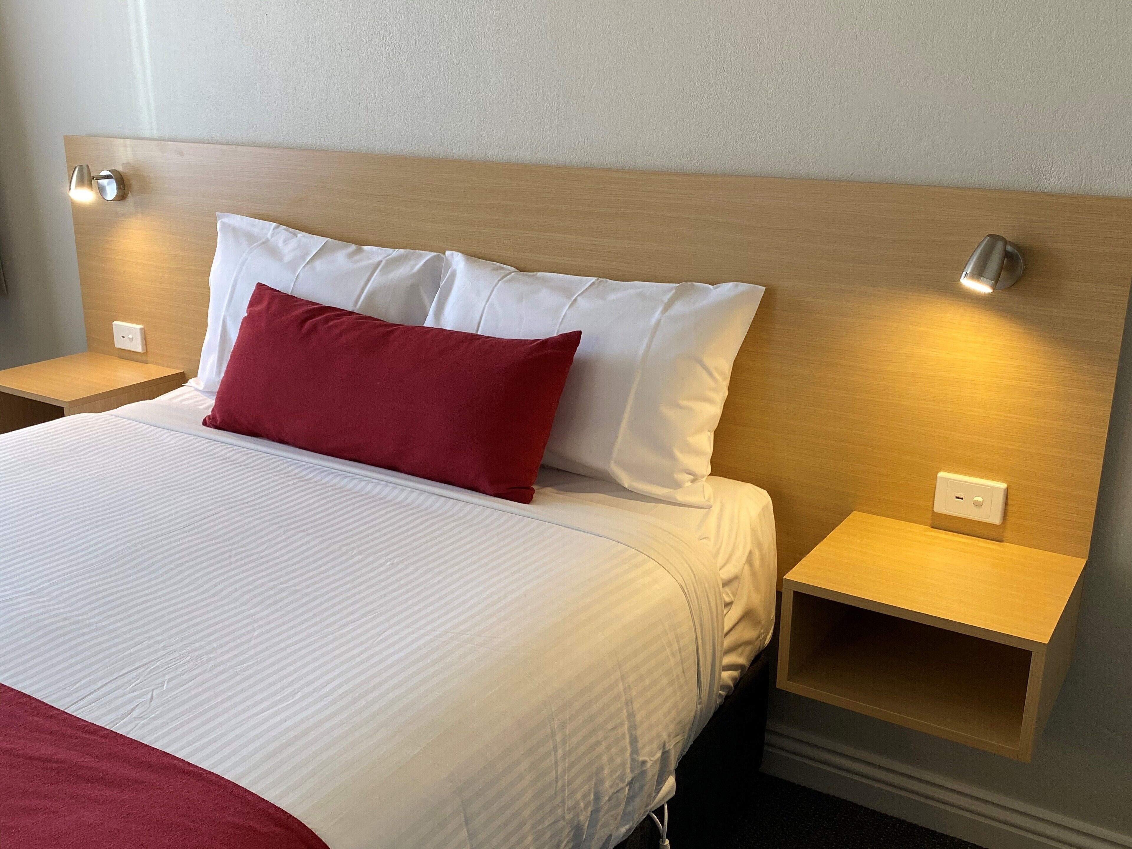 A neatly made bed with white and red pillows, wooden headboard, and bedside tables with wall-mounted lamps.
