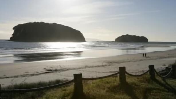 A beach with two large rock formations, a wooden fence, and a clear sky. - Marine Reserved Holiday Apartments (Whangamata)