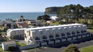 A white multi-story building with a row of balconies, located near a body of water and surrounded by greenery. - Marine Reserved Holiday Apartments (Whangamata)