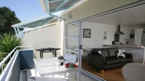 A modern balcony with a glass door leading to a living area. There is a dining table with chairs, a sofa, and a kitchen visible through the glass. - Marine Reserved Holiday Apartments (Whangamata)