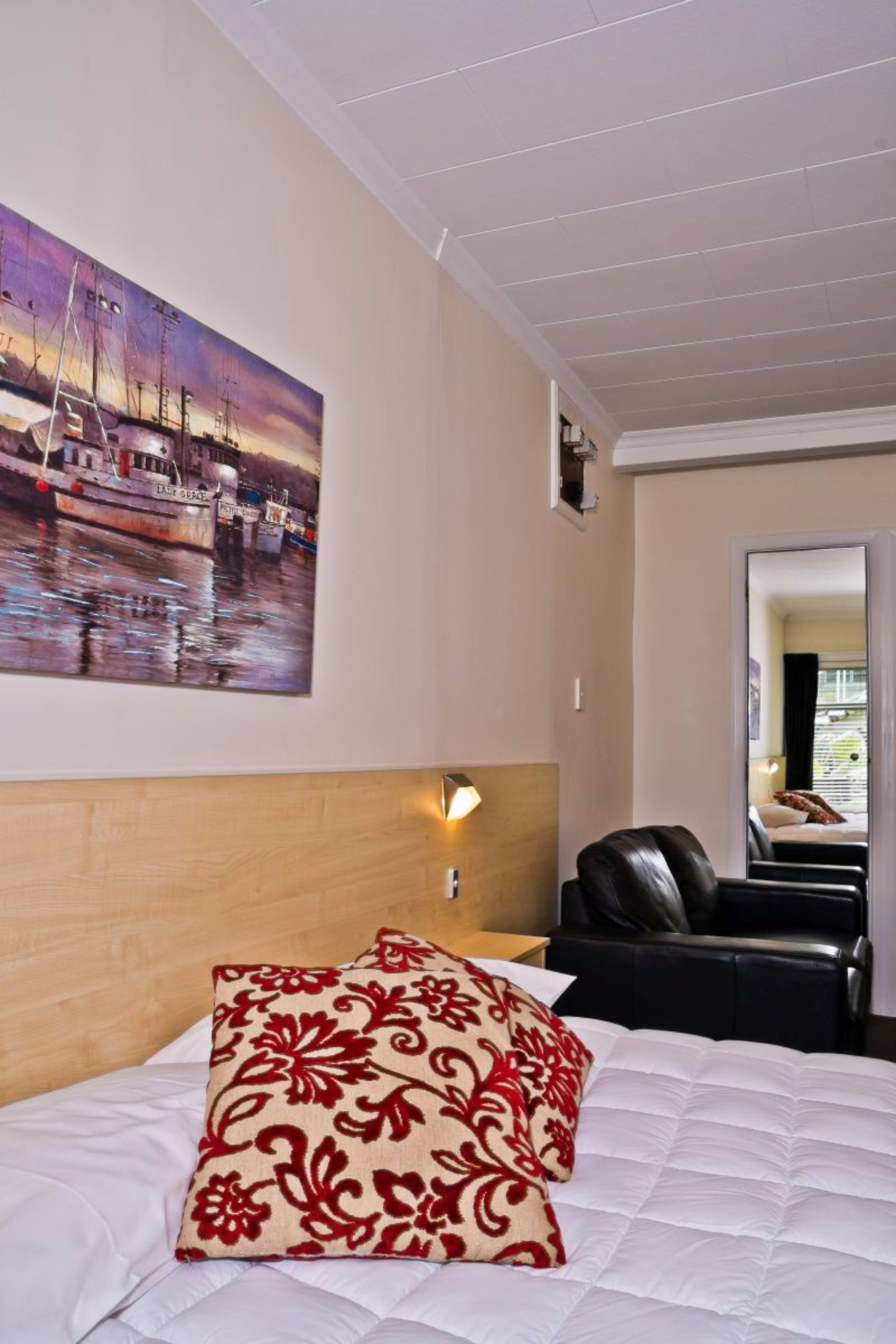 Habitación de hotel con cama, cabecera, cuadro de barcos, una silla y espejo.